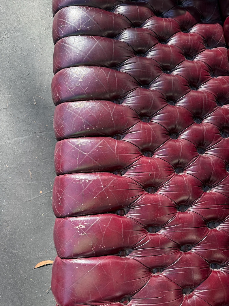 May include: Close-up of a burgundy leather upholstered surface with a tufted design. The leather shows signs of wear and tear, with visible scratches and indentations. The surface is likely part of a piece of furniture, such as a sofa or headboard.