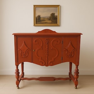 May include: A red-orange antique buffet with ornate carvings on the doors and sides. The buffet has four turned legs with brass casters and a decorative apron. A framed landscape painting hangs above the buffet.