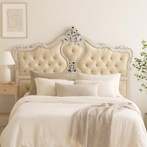 May include: A luxurious bedroom scene featuring a tufted headboard with ornate silver trim. The bed is adorned with cream-colored bedding, pillows, and a fringed throw blanket. A neutral rug and artwork complete the elegant, inviting space.