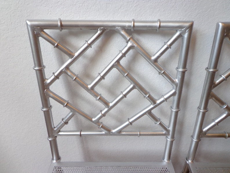 May include: Close-up of two silver metal chairs with a bamboo-style back design. The backs feature a geometric pattern with horizontal and diagonal bars, and the frames have a metallic finish. The chairs are suitable for indoor or outdoor use.