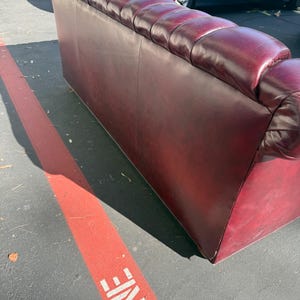 May include: A burgundy leather sofa with a tufted back and armrests. The sofa has a classic design and appears to be in good condition. The sofa is placed outside on a paved surface.