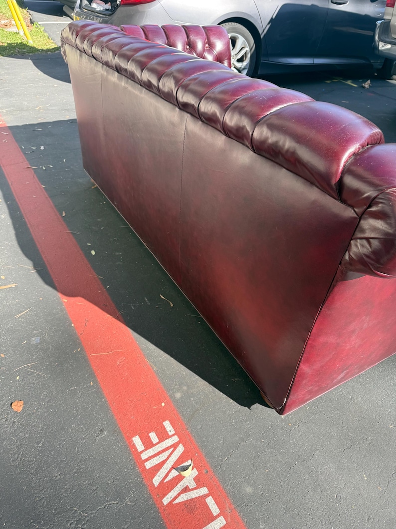 May include: A burgundy leather sofa with a tufted back and armrests. The sofa has a classic design and appears to be in good condition. The sofa is placed outside on a paved surface.