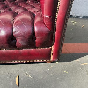 May include: Close-up of a burgundy leather armchair with button tufting. The armrest is detailed with brass-colored nailhead trim. The leather shows signs of wear, adding character to the vintage furniture.