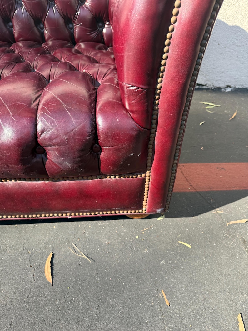 May include: Close-up of a burgundy leather armchair with button tufting. The armrest is detailed with brass-colored nailhead trim. The leather shows signs of wear, adding character to the vintage furniture.