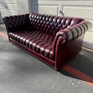 May include: A burgundy leather Chesterfield sofa with a button-tufted back and seat. The sofa has rolled arms and a low profile, with nailhead trim along the base and arms. The sofa is a classic piece of furniture.