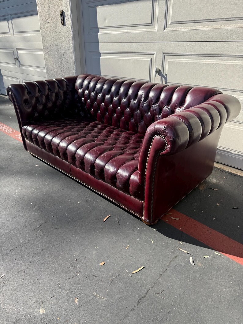 May include: A burgundy leather Chesterfield sofa with a button-tufted back and seat. The sofa has rolled arms and a low profile, with nailhead trim along the base and arms. The sofa is a classic piece of furniture.