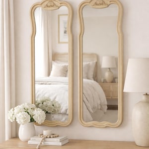 May include: Two tall, cream-colored decorative mirrors with ornate tops are displayed on a light wood console table. A white vase of flowers, books, and a beaded garland adorn the table. The mirrors reflect a bedroom scene.