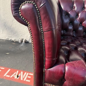 May include: Close-up of a burgundy leather Chesterfield sofa arm. The armrest is tufted with a row of brass nailhead trim. The sofa has a classic, elegant design.
