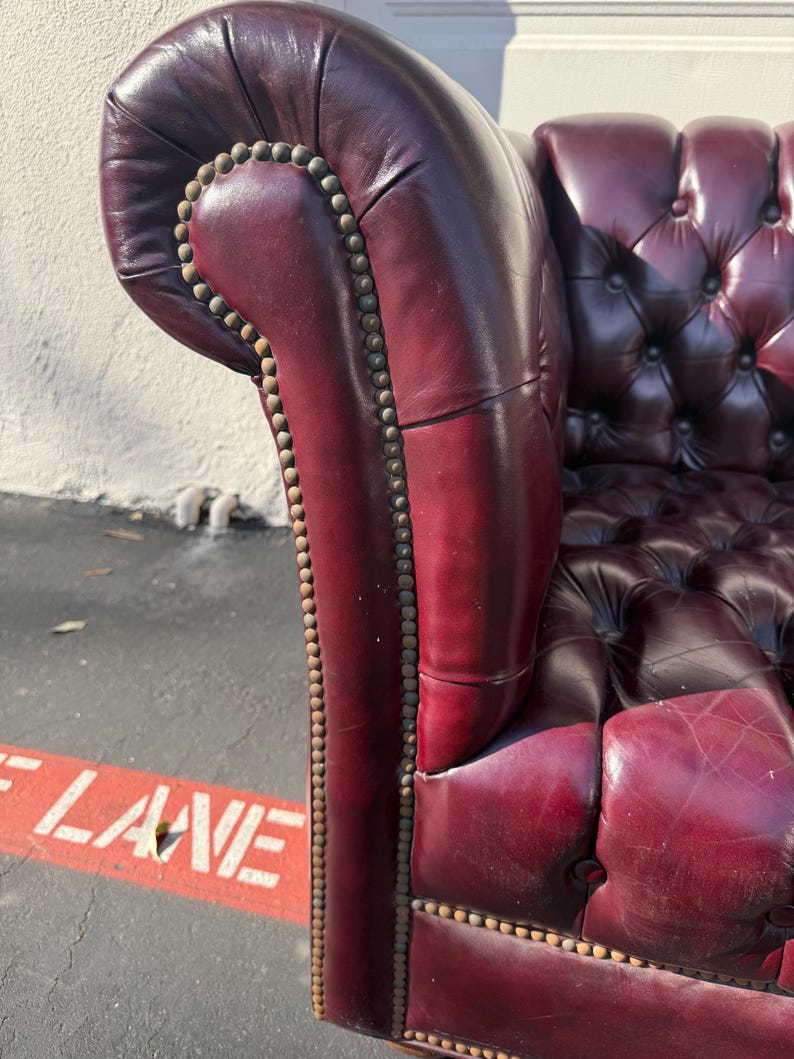 May include: Close-up of a burgundy leather Chesterfield sofa arm. The armrest is tufted with a row of brass nailhead trim. The sofa has a classic, elegant design.