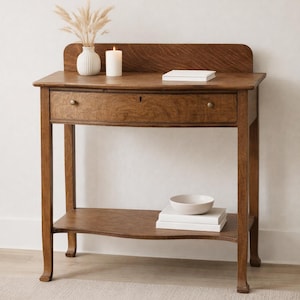 May include: A wooden console table with a drawer and a lower shelf. A white vase with dried plants, a lit candle, a stack of books, and a small bowl are on the table. The table is brown and has a curved back.