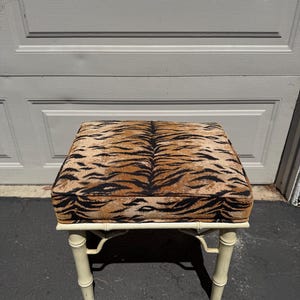 May include: A square ottoman with a tiger stripe pattern on the cushion. The frame is painted a cream color and has a bamboo-style design. The ottoman is a decorative furniture piece.