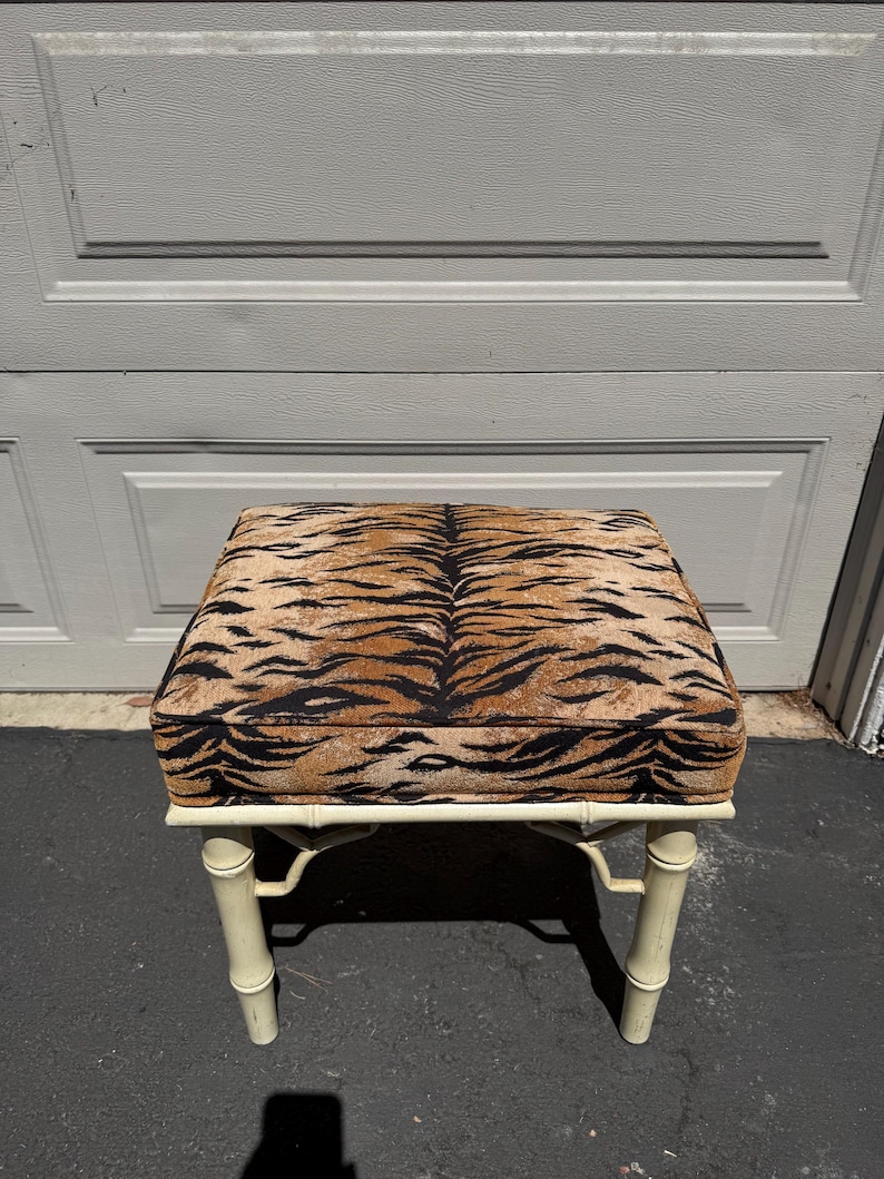 May include: A square ottoman with a tiger stripe pattern on the cushion. The frame is painted a cream color and has a bamboo-style design. The ottoman is a decorative furniture piece.