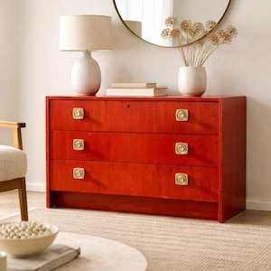 May include: A red three-drawer dresser with brass knobs. A white lamp with a beige lampshade sits on top, along with books and a white vase with dried flowers. A round mirror hangs on the wall.