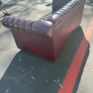 May include: A burgundy leather Chesterfield sofa with a tufted back and rolled arms. The sofa has a classic design and is made of a rich, dark red-brown leather. The sofa is placed outdoors.