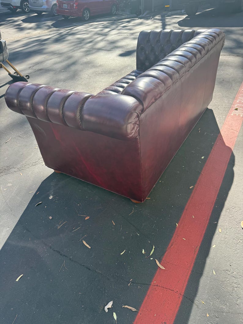 May include: A burgundy leather Chesterfield sofa with a tufted back and rolled arms. The sofa has a classic design and is made of a rich, dark red-brown leather. The sofa is placed outdoors.