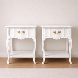 May include: Two white bedside tables with a classic design. Each table features a single drawer with a gold-colored ornate handle, a lower shelf, and curved legs. The tables are set against a neutral wall.