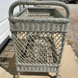 May include: A gray wicker serving cart with a handle and wheels. The cart has two shelves and is made of woven wicker.