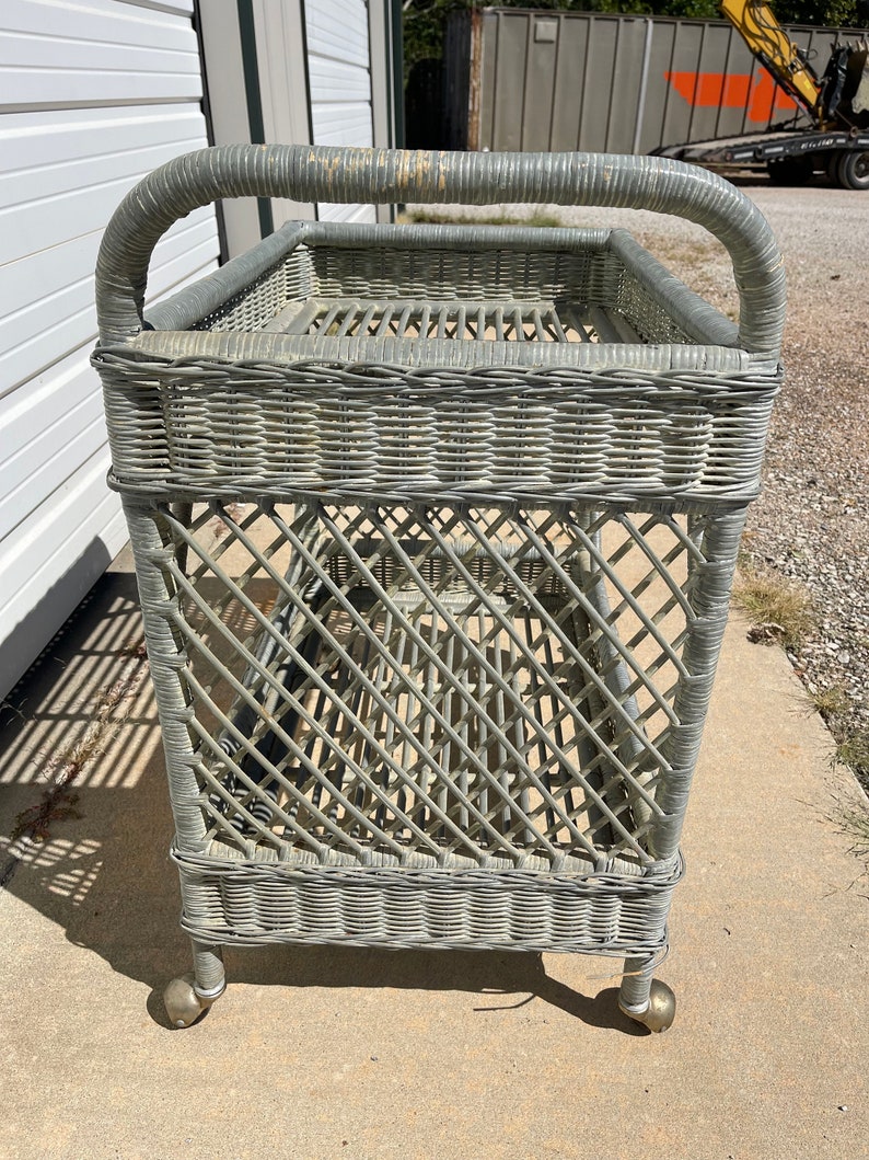 May include: A gray wicker serving cart with a handle and wheels. The cart has two shelves and is made of woven wicker.
