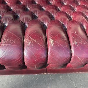 May include: Close-up of a burgundy leather sofa with a tufted design. The leather shows signs of wear, with visible scratches and discoloration. The sofa's cushions are rounded and well-defined, suggesting a classic style.