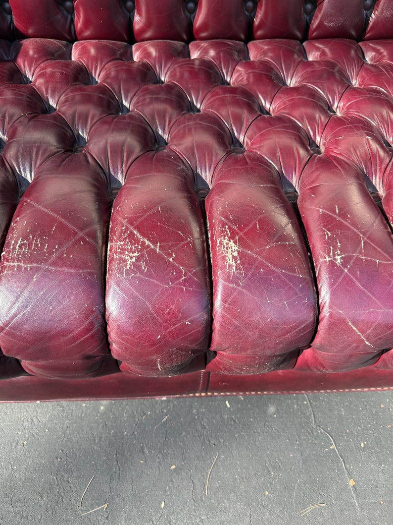 May include: Close-up of a burgundy leather sofa with a tufted design. The leather shows signs of wear, with visible scratches and discoloration. The sofa's cushions are rounded and well-defined, suggesting a classic style.