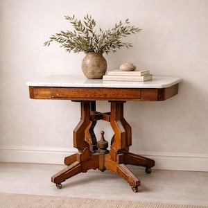 May include: An antique wooden side table with a white marble top. The table has a drawer and ornate wooden legs with small wheels. A vase with greenery, a stack of books, and a stone are on the table. The table is approximately 30 inches tall.