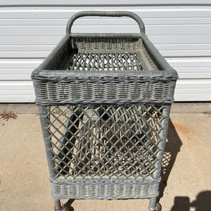 May include: A gray wicker serving cart with a handle and wheels. The cart has a woven design and is made of natural materials.
