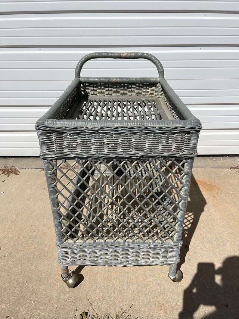 May include: A gray wicker serving cart with a handle and wheels. The cart has a woven design and is made of natural materials.