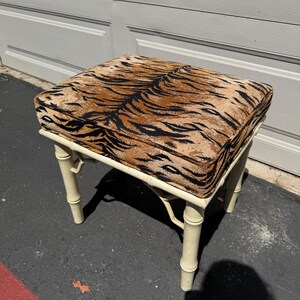 May include: A square, cream-colored bamboo-style stool with a tiger print cushion. The cushion features a brown and black pattern. The stool has four legs and is suitable for use as a footstool or accent piece.