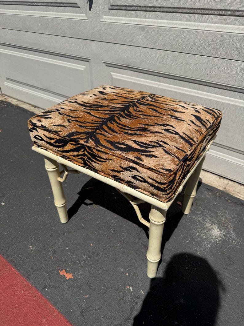 May include: A square, cream-colored bamboo-style stool with a tiger print cushion. The cushion features a brown and black pattern. The stool has four legs and is suitable for use as a footstool or accent piece.