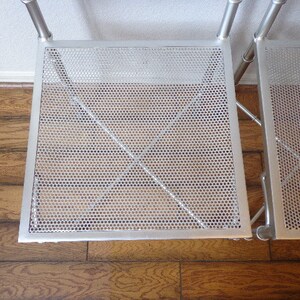 May include: A silver metal shelving unit with two perforated square shelves. The shelves have a grid-like pattern of small holes and are supported by tubular metal legs and crossbars. The unit is set against a white wall and a wooden floor.
