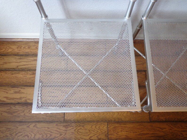 May include: A silver metal shelving unit with two perforated square shelves. The shelves have a grid-like pattern of small holes and are supported by tubular metal legs and crossbars. The unit is set against a white wall and a wooden floor.
