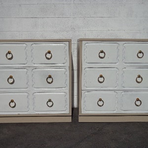 May include: Two white dresser chests with six drawers each. The drawers have decorative raised edges and gold-colored ring pulls. The chests are sitting on a white tiled floor.