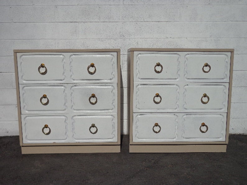 May include: Two white dresser chests with six drawers each. The drawers have decorative raised edges and gold-colored ring pulls. The chests are sitting on a white tiled floor.