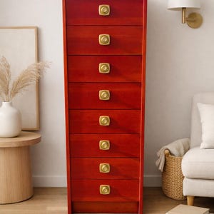 May include: A tall, red wooden dresser with seven drawers, each featuring a round, gold-colored knob. The dresser is set against a white wall, with a neutral-toned rug and a side table with a vase.