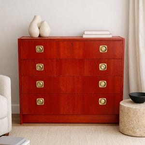 May include: A red wooden dresser with four drawers and brass hardware. Two white vases and a stack of books sit on top. The dresser is in a room with a neutral rug and a white curtain.
