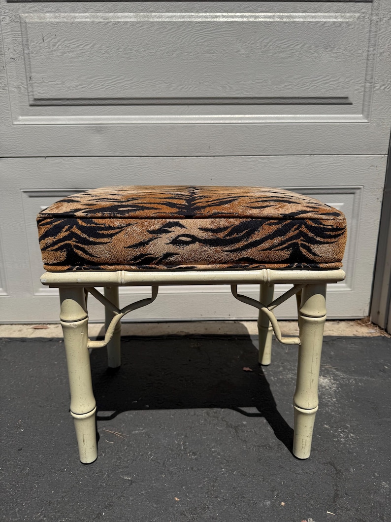 May include: A small, cream-colored bamboo-style stool with a rectangular seat. The seat is upholstered with a brown and black tiger stripe pattern. The stool has four legs and is set against a neutral background.