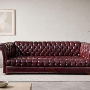 May include: A burgundy leather Chesterfield sofa with button tufting. The sofa has rolled arms and a low profile. The sofa is placed on a light gray rug in a room with neutral walls and decor. A framed artwork and a side table with a lamp are visible.
