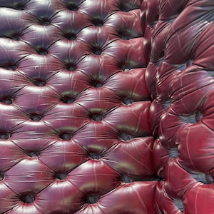 May include: Close-up of a section of a burgundy leather upholstered surface. The surface is tufted with a diamond pattern, and the buttons are dark. The leather has a slight sheen, and the overall effect is luxurious.