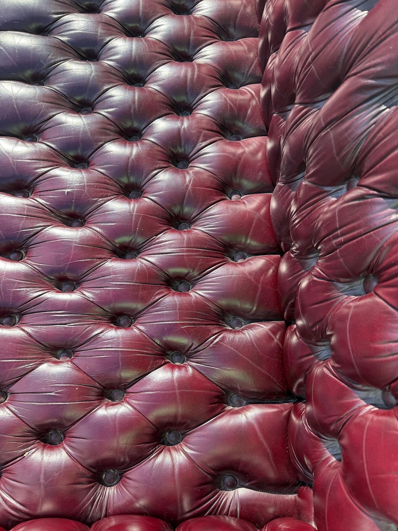 May include: Close-up of a section of a burgundy leather upholstered surface. The surface is tufted with a diamond pattern, and the buttons are dark. The leather has a slight sheen, and the overall effect is luxurious.