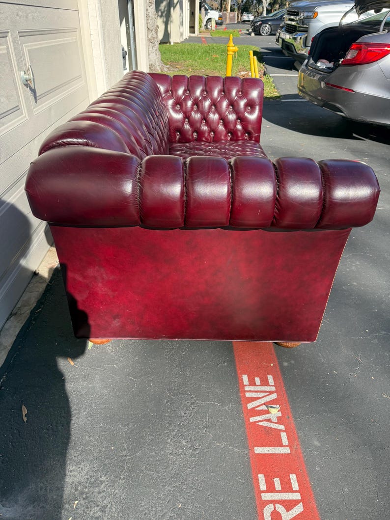May include: A burgundy leather Chesterfield sofa with a tufted back and rolled arms. The sofa has a low profile and is set on wooden feet. The sofa is a classic piece of furniture.