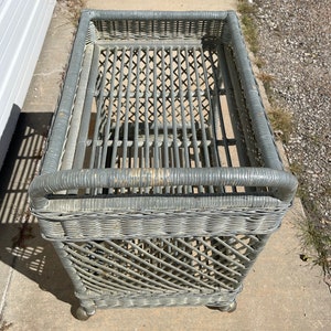 May include: A gray wicker rolling cart with a woven design. The cart has a rectangular top and a bottom shelf. The cart is on wheels.
