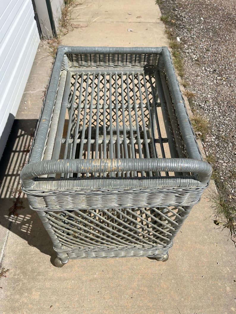 May include: A gray wicker rolling cart with a woven design. The cart has a rectangular top and a bottom shelf. The cart is on wheels.