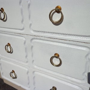 May include: White dresser with four drawers and gold ring-shaped handles. The dresser has a decorative, scalloped edge.