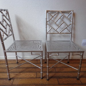 May include: Two silver chairs with bamboo-style frames and woven seats. The chairs have a lattice design on the backrests. They are set against a neutral wall and wooden floor.