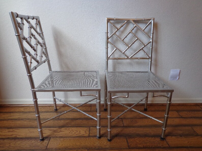 May include: Two silver chairs with bamboo-style frames and woven seats. The chairs have a lattice design on the backrests. They are set against a neutral wall and wooden floor.