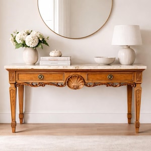 May include: A wooden console table with a marble top, two drawers, and ornate carvings. A round mirror hangs above the table, which is decorated with a lamp, vase of flowers, books, and a bowl. The table is approximately 60 inches long.