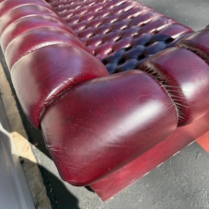 May include: A burgundy leather sofa with a button-tufted back and seat. The sofa has rounded arms and a low profile. The leather has a glossy finish, and the sofa appears to be well-maintained. The sofa is a classic piece of furniture.