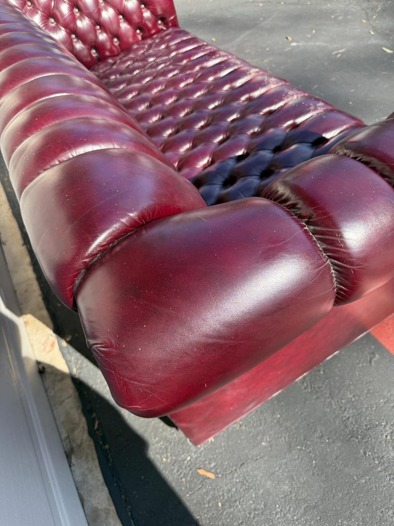May include: A burgundy leather sofa with a button-tufted back and seat. The sofa has rounded arms and a low profile. The leather has a glossy finish, and the sofa appears to be well-maintained. The sofa is a classic piece of furniture.