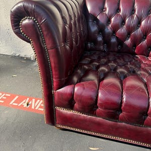 May include: A burgundy leather Chesterfield sofa with button tufting and brass nailhead trim. The armrest curves outward, and the sofa appears to be well-worn. The sofa is a classic piece of furniture.