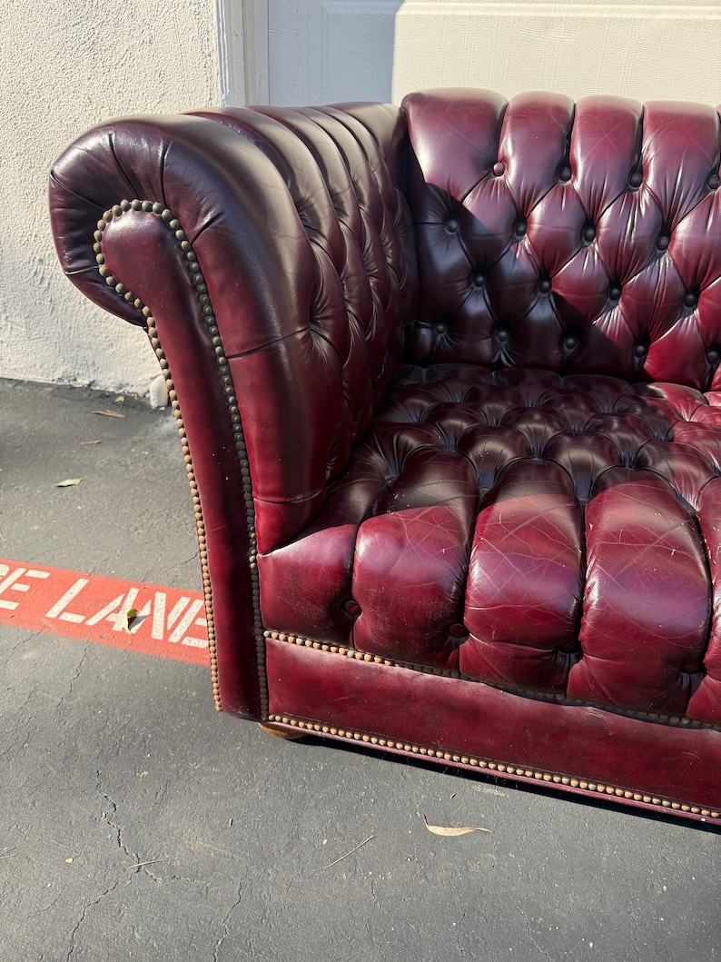 May include: A burgundy leather Chesterfield sofa with button tufting and brass nailhead trim. The armrest curves outward, and the sofa appears to be well-worn. The sofa is a classic piece of furniture.